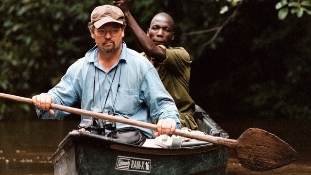 A white man sits up front of a canoe with cap and open collar with an African man behind, both paddling.