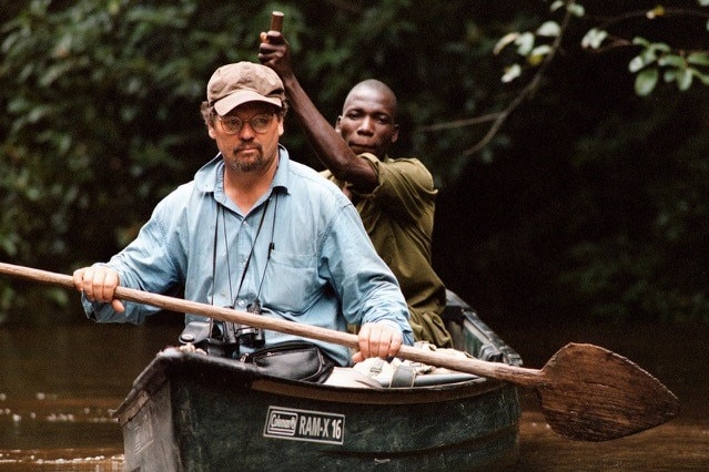 A white man sits up front of a canoe with cap and open collar with an African man behind, both paddling.