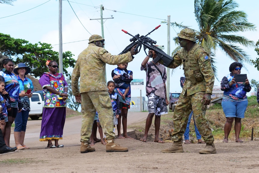 Men in uniform cross guns. Islanders stand around.