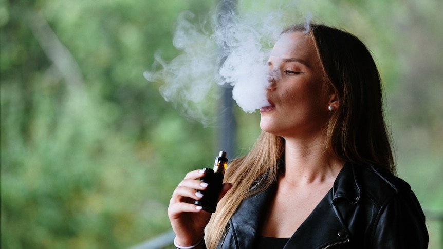 A close-up of a woman with long brown hair vaping, blowing out a big cloud of smoke.