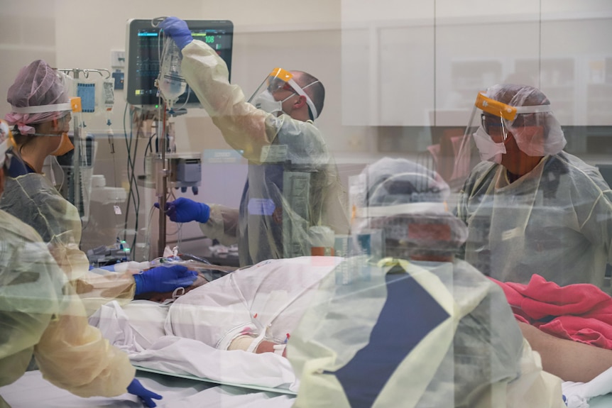 A shot through the glass window showing health staff clad in PPE working on a patient surrounded by machines.