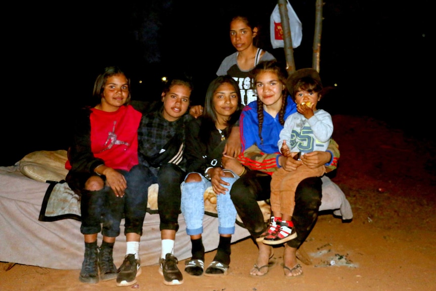 A group of Aboriginal teenage girls, one holding a young boy, sitting on a camp bed at night.