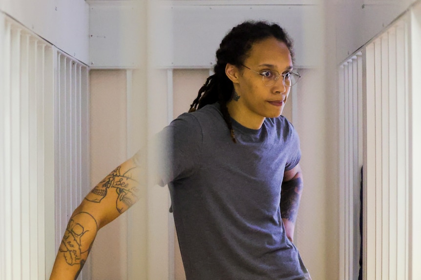 Una mujer negra alta con gafas de montura metálica y una camiseta gris se encuentra en una venta de prisión