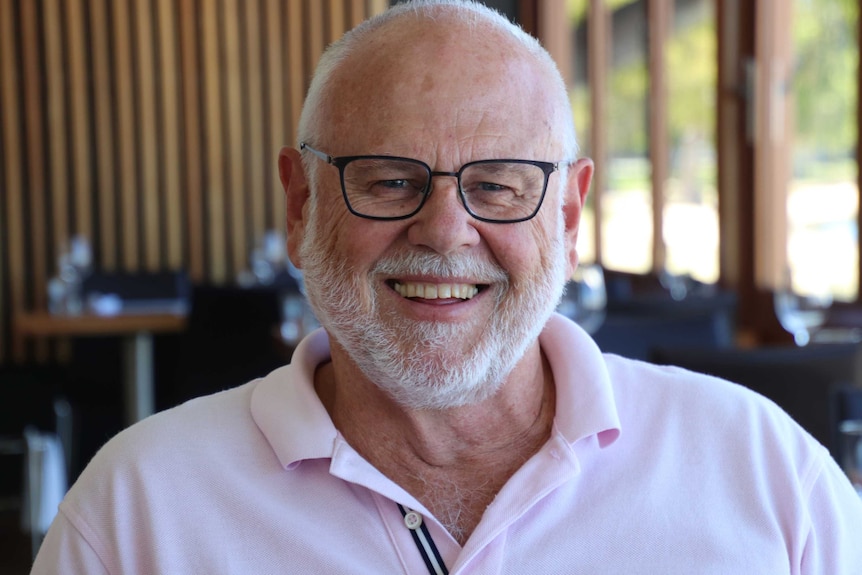 A head and shoulder shot of a smiling older man with a bald heard, white beard and black spectacles, wearing a pink shirt.