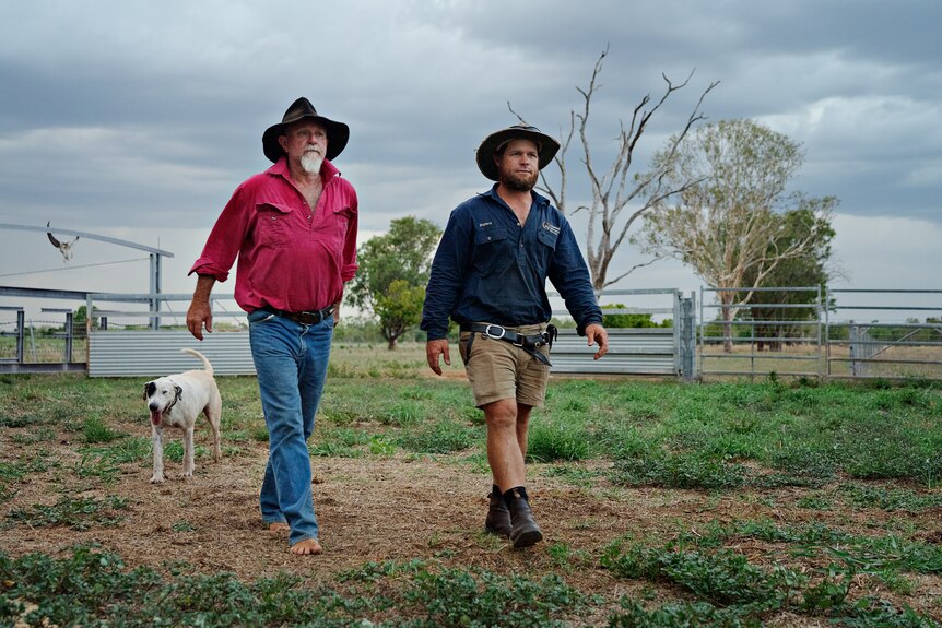 Two men walking across a farm paddock with a dog behind them.