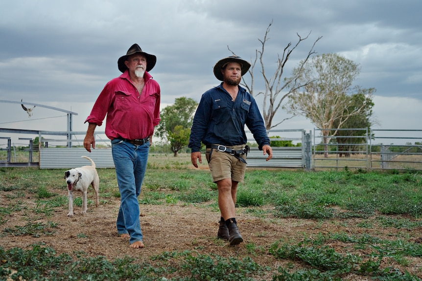 Two men walking across a farm paddock with a dog behind them.