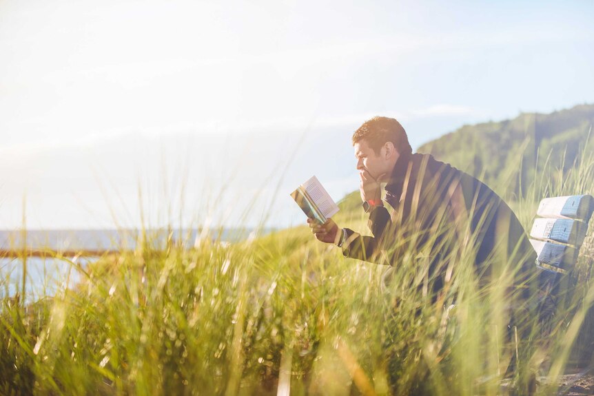 Man sitting on a bench reading a book, surrounded by long grass