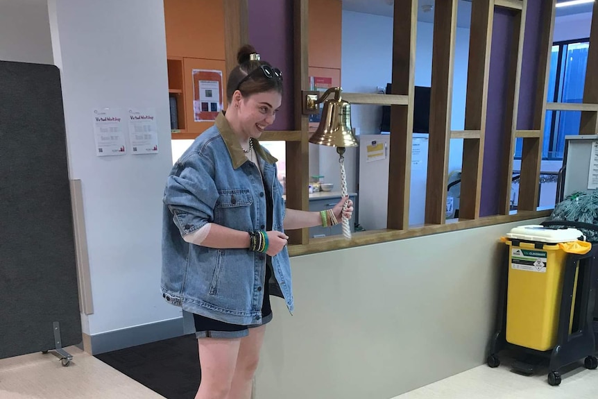 A young woman in a denim jacket rings a gold bell in a hopsital ward, she looks happy