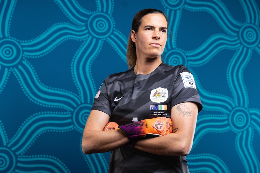 Matildas goalie Lydia Williams poses for a portrait in a black jersey. She is looking into the distance with her arms crossed.
