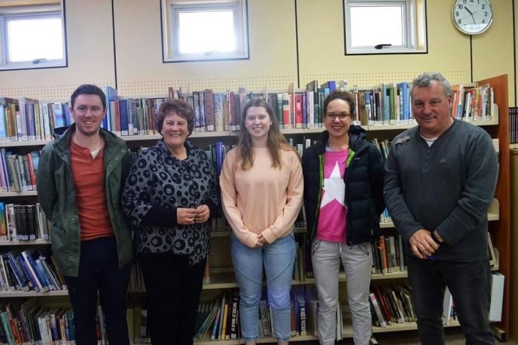 Five adults stand in a library and they are all smiling.  There are many books behind them.