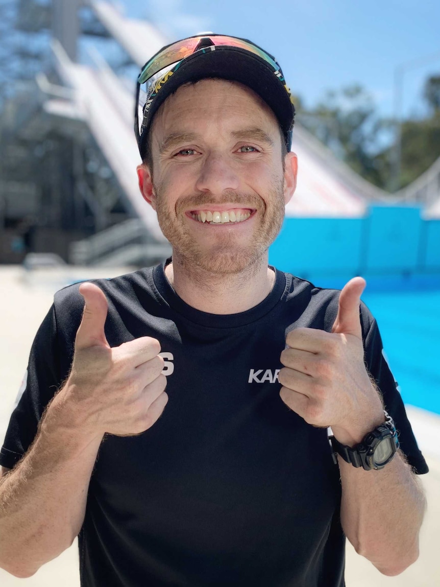 A young man with light beard in sports attire and black cap smiles and gives thumbs up in pool area.