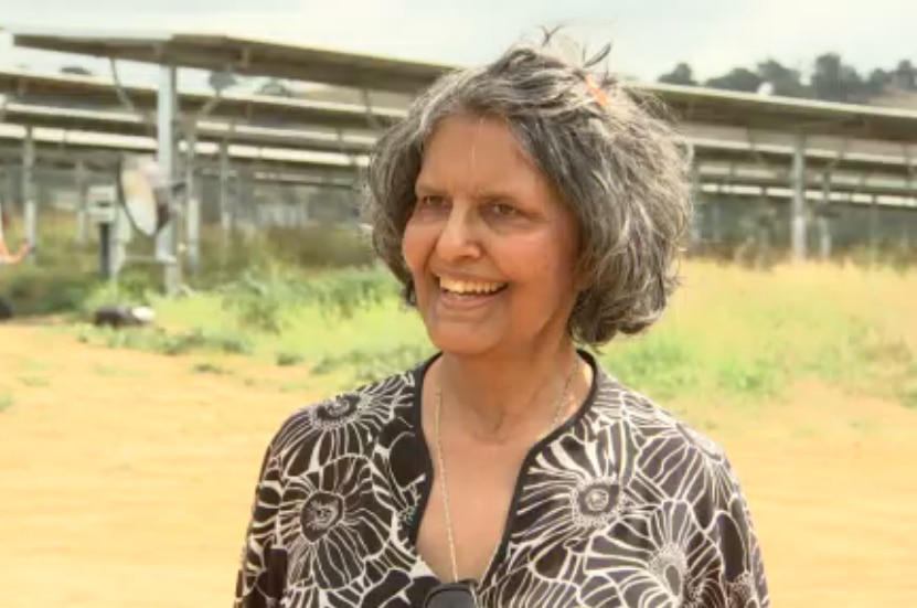 A woman smiles passed the camera while standing in a field with solar panels behind her