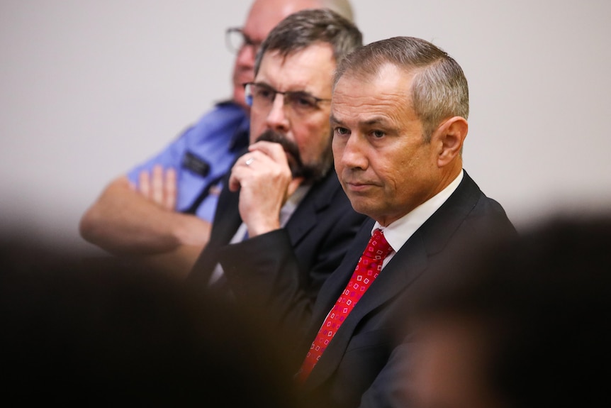 Roger Cook watches a press conference.