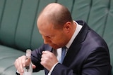 Josh Frydenberg coughs into his hand. He's holding a bottle of water in the other hand while standing in the parliament