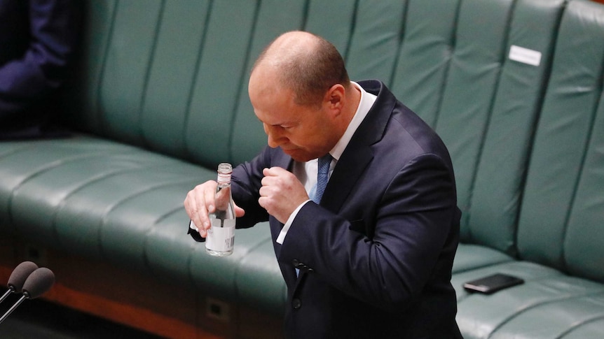 Josh Frydenberg coughs into his hand. He's holding a bottle of water in the other hand while standing in the parliament