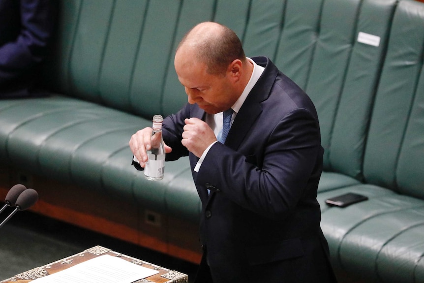 Josh Frydenberg coughs into his hand. He's holding a bottle of water in the other hand while standing in the parliament