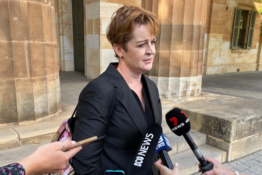 A woman with short reddy-brown hair speaks to reporters with microphones