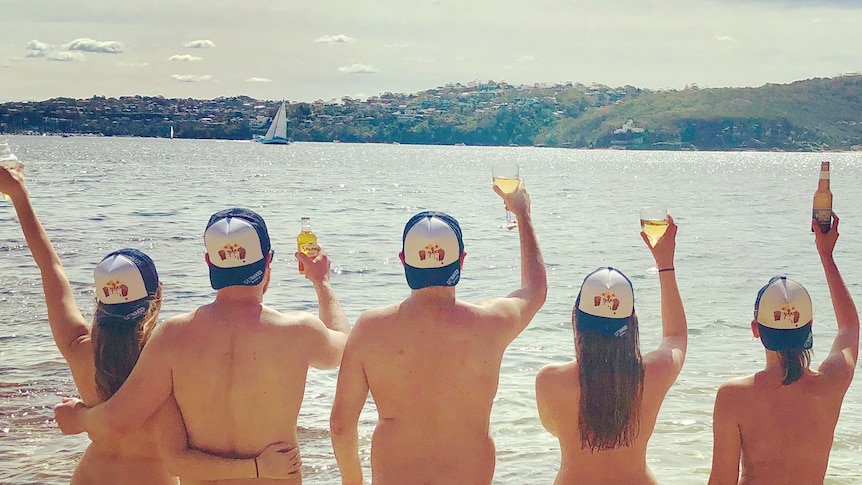 A group of naked people raise their glasses while standing on a beach and facing towards the water