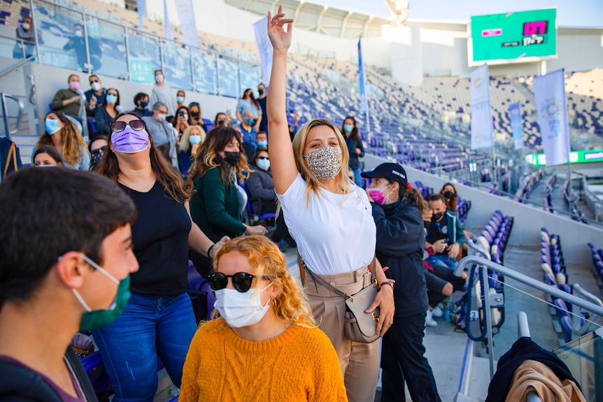 A young woman in a face mask cheering in the stadium surrounded by other people in face masks