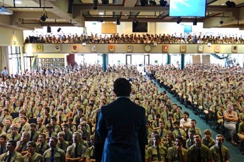 A photo of the back of a man standing at a lectern in front of a crowd of people.