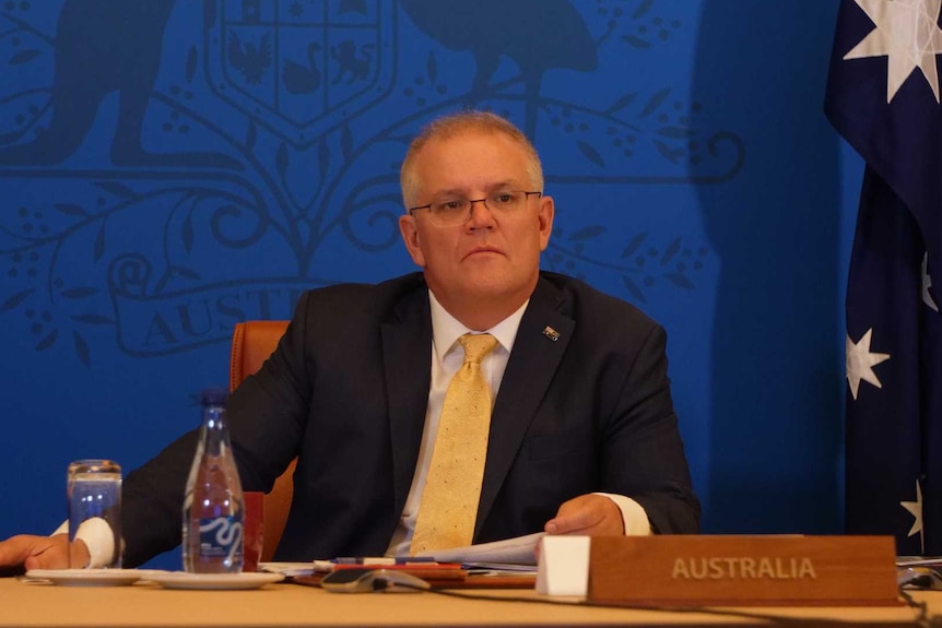 Scott Morrison looks slightly to his left as he sits in front of a blue wall with flags to his left and right.