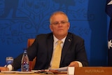 Scott Morrison looks slightly to his left as he sits in front of a blue wall with flags to his left and right.