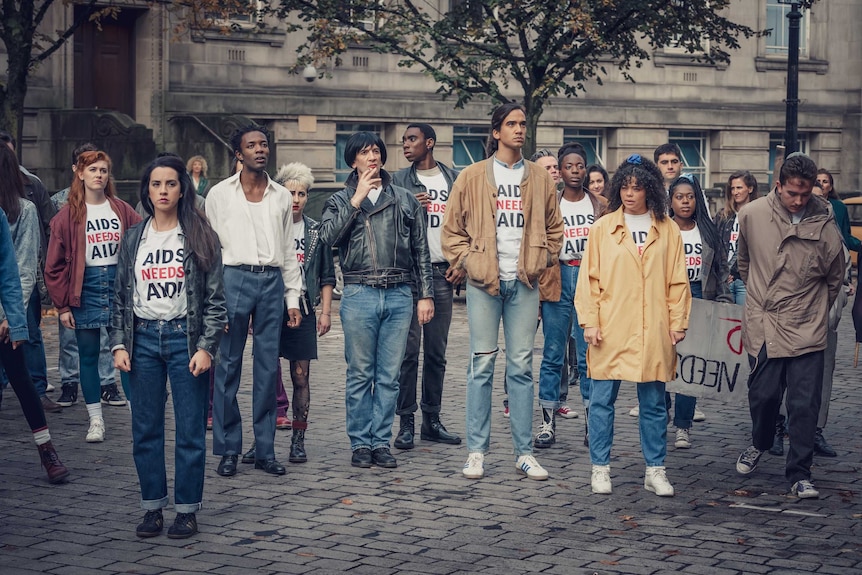 A still from the miniseries It's a Sin with a group of protestors on a London street wearing tshirts that say "AIDS NEEDS AID"