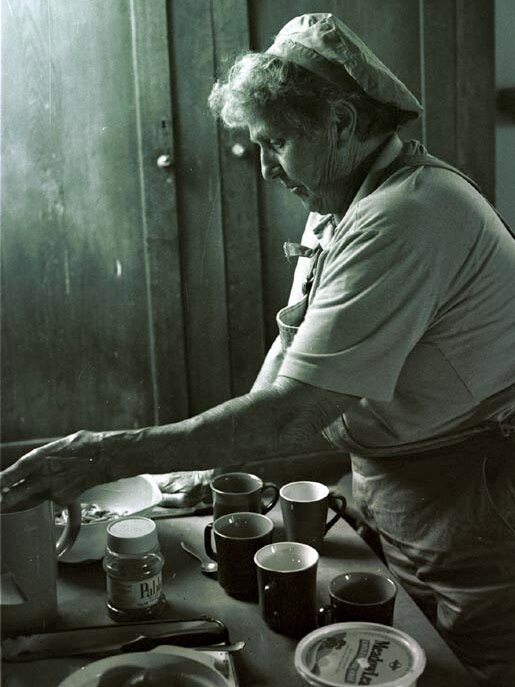 Older lady, in apron, makes cups of tea at a table inside the shack.