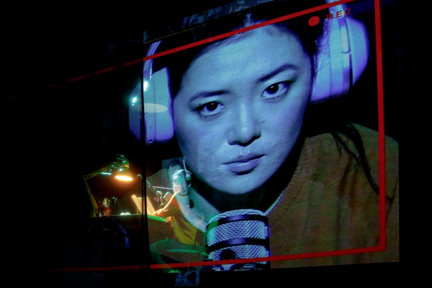 A woman in headphones is hunched over a microphone at a table, with a close-up image of her face projected on a screen behind 