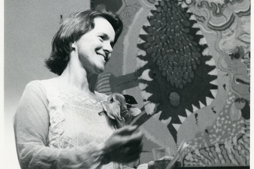 Binns, in a dress with a flower pinned to her lapel, stands smiling in front of a painting of a vagina with teeth.