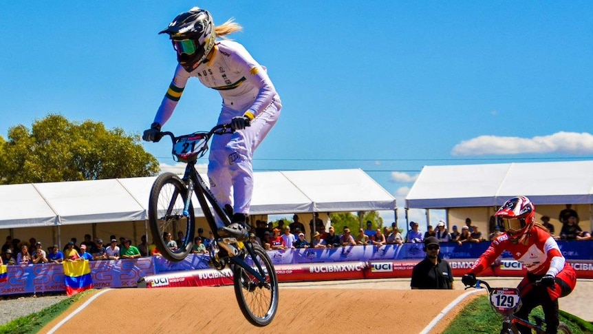 A BMX rider in white race suit and black helmet goes over a jump with both wheels in the air
