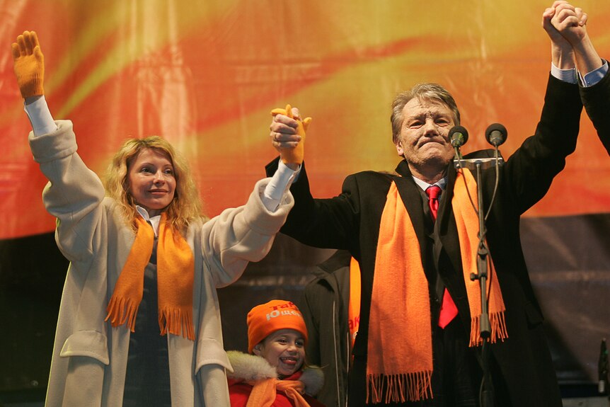 a woman and a man wearing orange scarfs hold each others' hands up in front of an orange sign at a rally