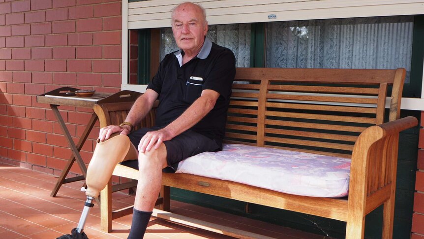 A man with a lower leg prosthesis is sitting on a bench out the front of his house. He is wearing a dark shirt and shorts.