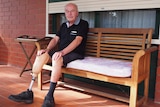 A man with a lower leg prosthesis is sitting on a bench out the front of his house. He is wearing a dark shirt and shorts.
