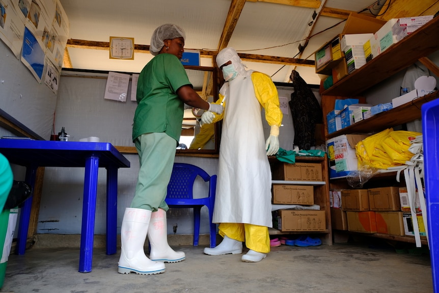 A person in green scrubs assists another person to dress in a yellow and white plastic protective outfit.