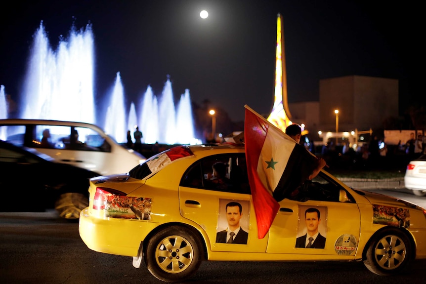 A boy holds a syrian flag out the window of a car featuring pictures of President Bashar Assad.