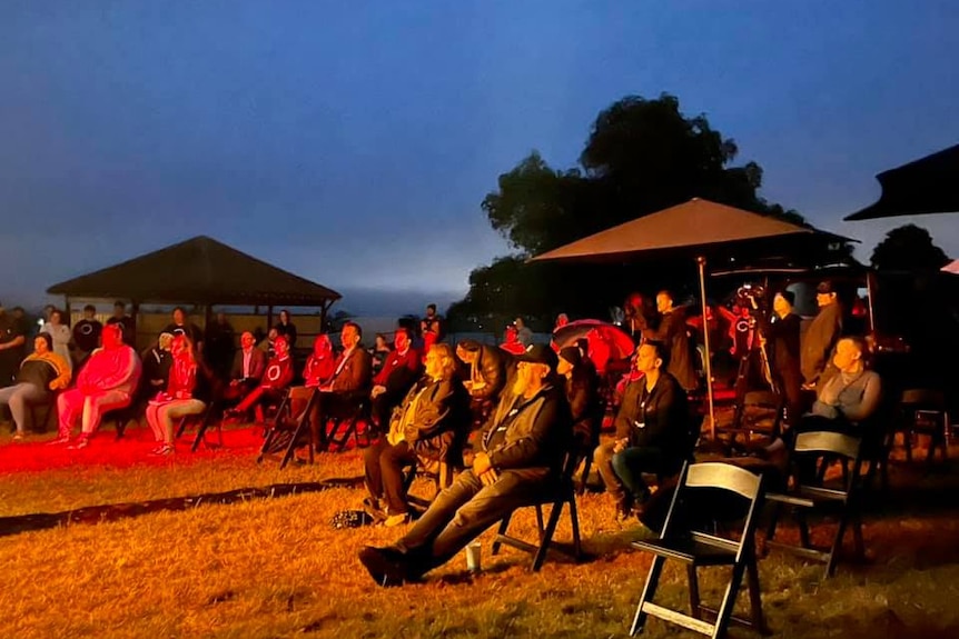 A group of people sit on chairs by Lake Wendouree, bathed in red and yellow light as they watch a dawn service.