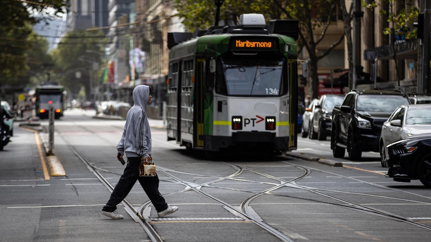 A man in a hoodie crosses a street in front of a tram in Melbourne's CBD.