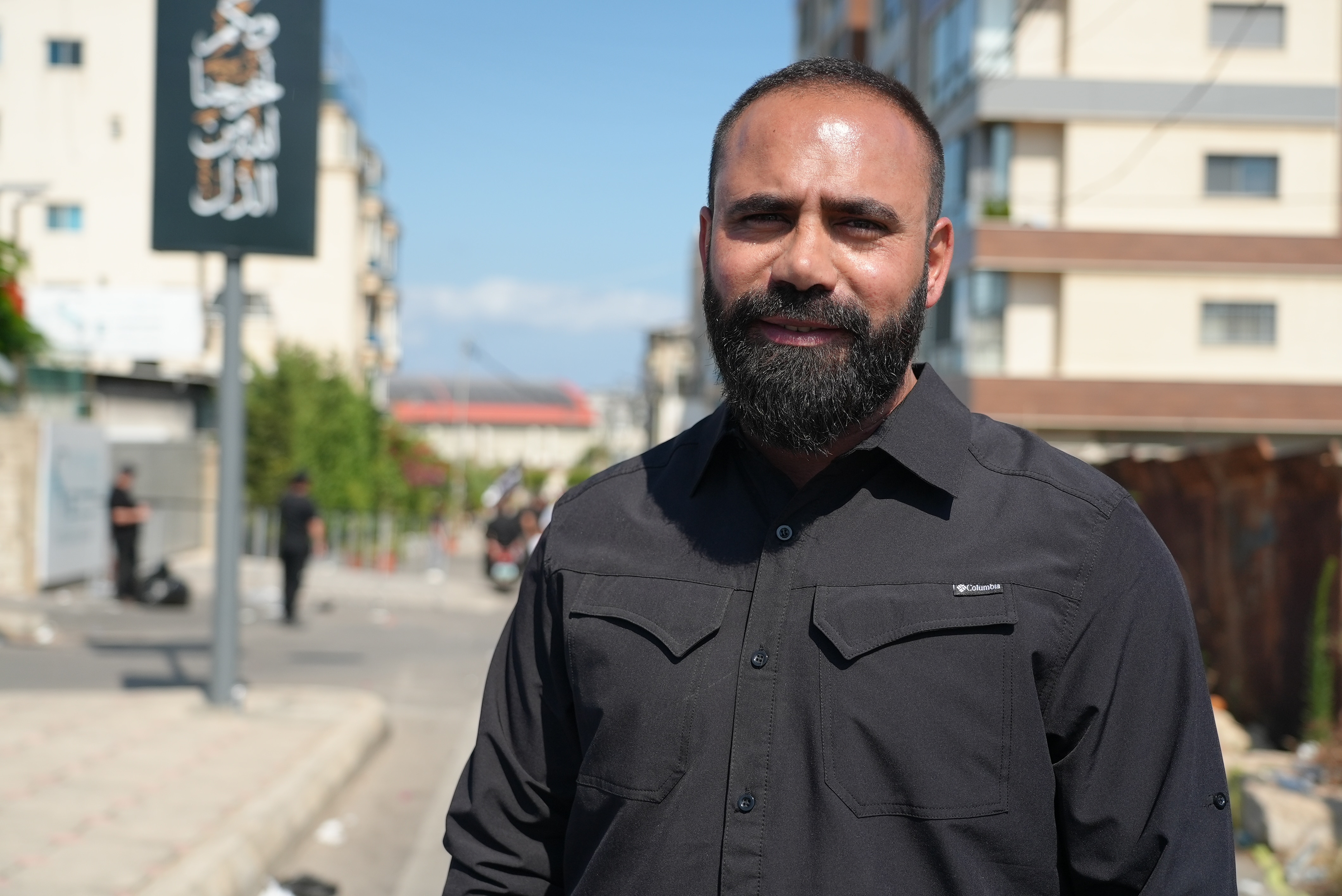 A man in his 30s, wearing a black shirt, has a thick black beard and is starting to bald.
