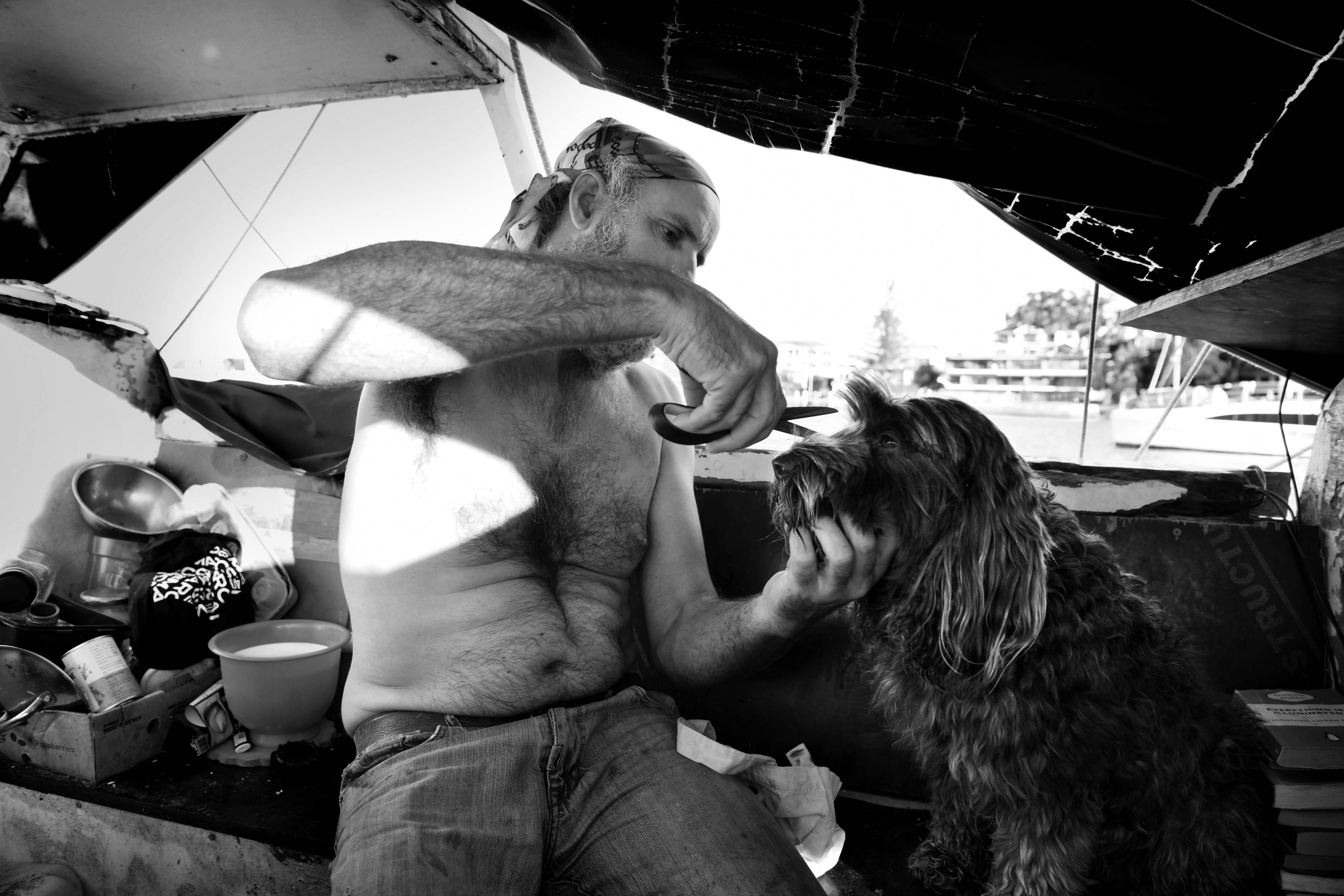 A shirtless man trims his dog's fur near its face with a pair of scissors