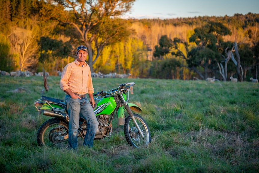 a man standing next to a farm bike in paddock with sheep in the background. 