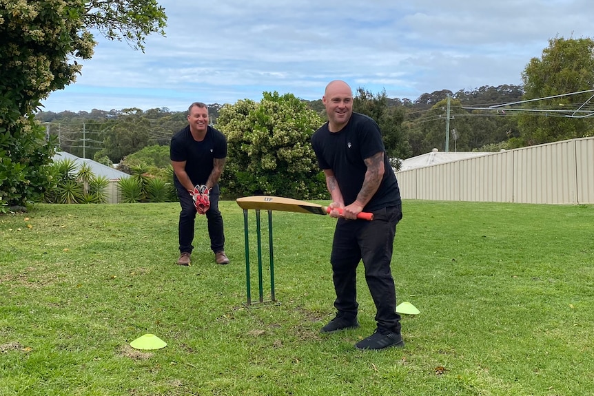 Two men dressed in black play cricket in a large grassy backyard, fence on the right, partly clouded blue sky.