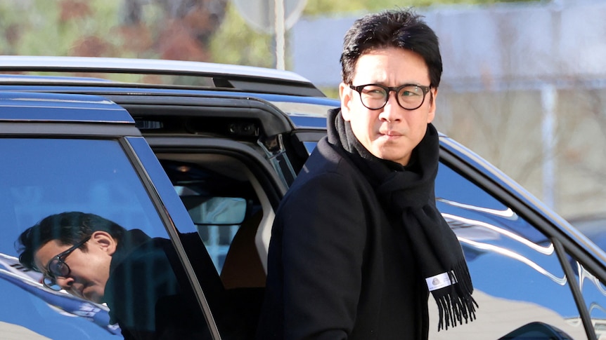 Lee Sun-kyun wears dark winter clothes as he exits the front right side of a car and looks over his right shoulder.