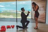 Brandon Jackson proposes on bended knee to Lara Markham in a red dress with a coastal view out the window.
