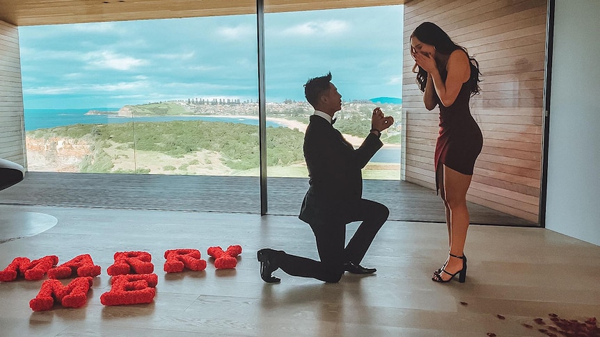 Brandon Jackson proposes on bended knee to Lara Markham in a red dress with a coastal view out the window.