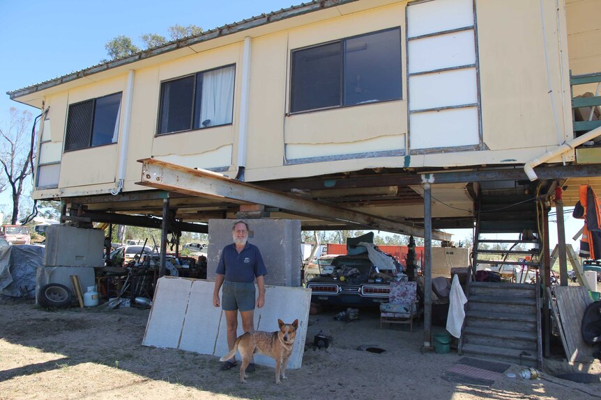 A man stands under his house which is on stilts next to a dog