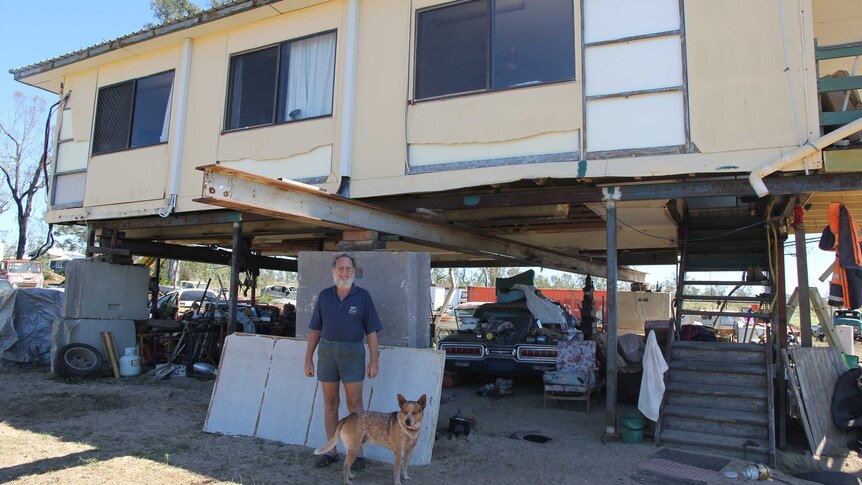 A man stands under his house which is on stilts next to a dog