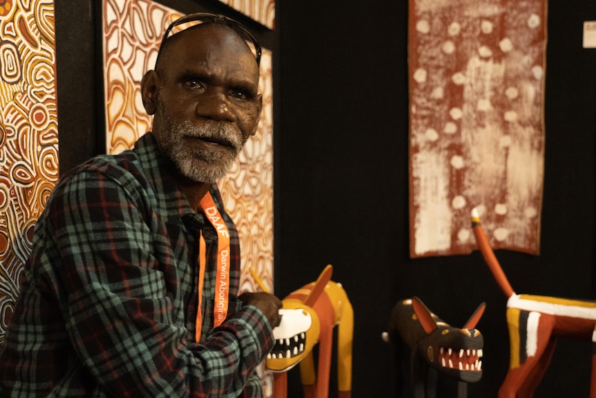 A man in a plaid shirt sitting down in front of a series of brightly colored paintings and sculptures on display in a gallery.