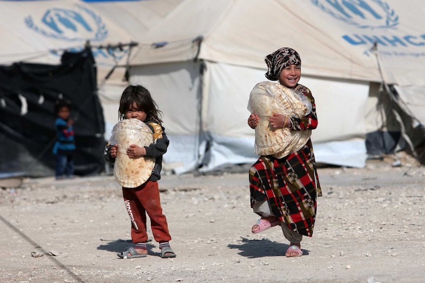 Two little girls carrying flat breads past UNHCR tents