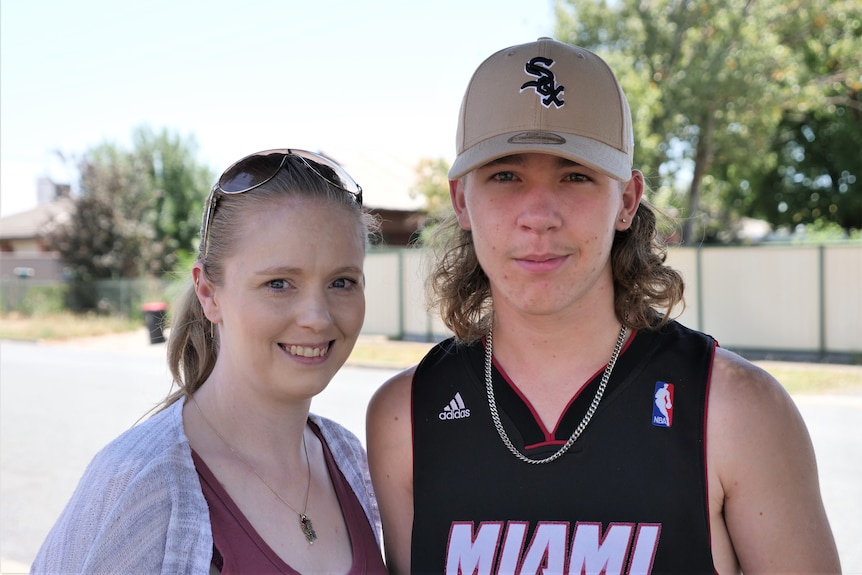 A woman with mid-length blonde hair stands next to a tall teenager with long curly hair who is wearing a baseball cap